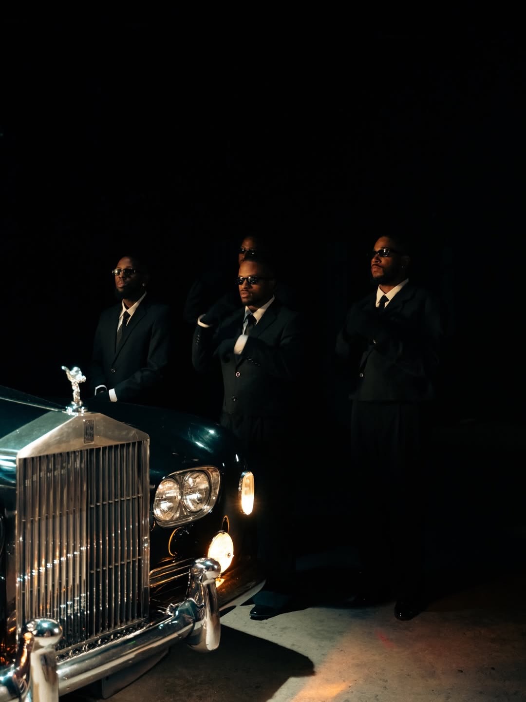 Three men beside the front of a classic Rolls-Royce on a dark set.